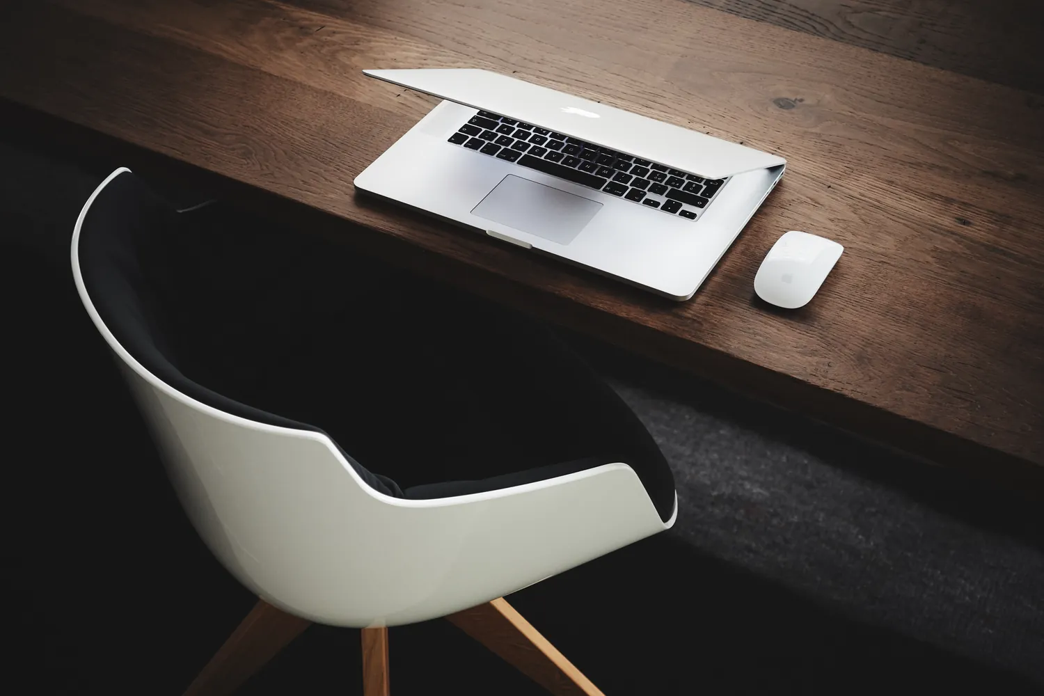 A laptop and mouse on a dark wooden desk beside a white chair