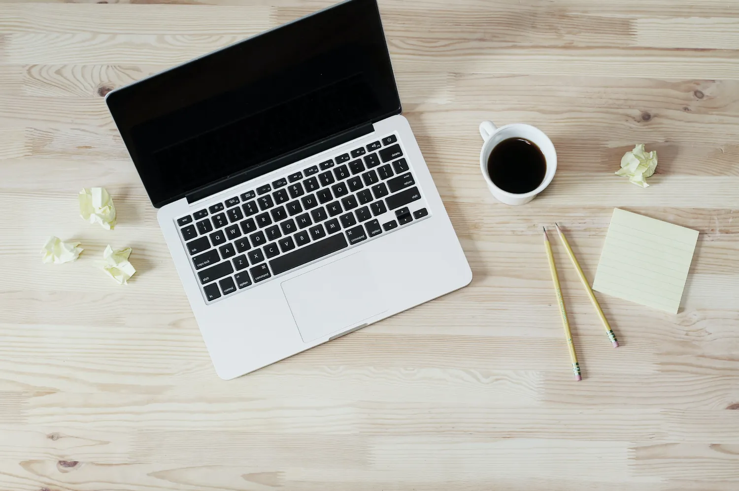 A laptop, a cup of coffee and a notepad on a pale wooden desk