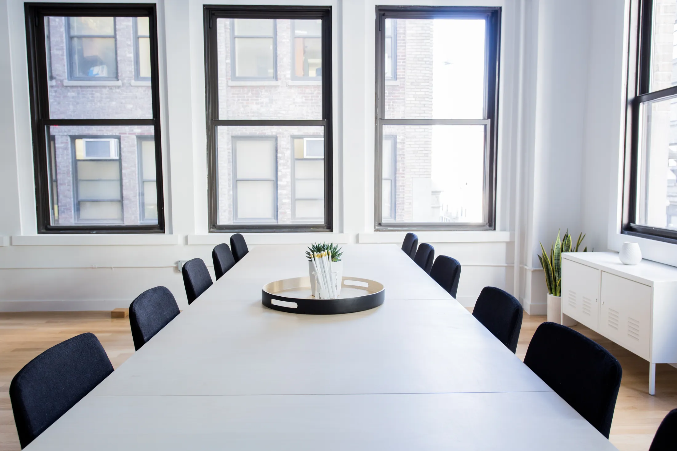 A long white meeting table with black chairs below tall windows