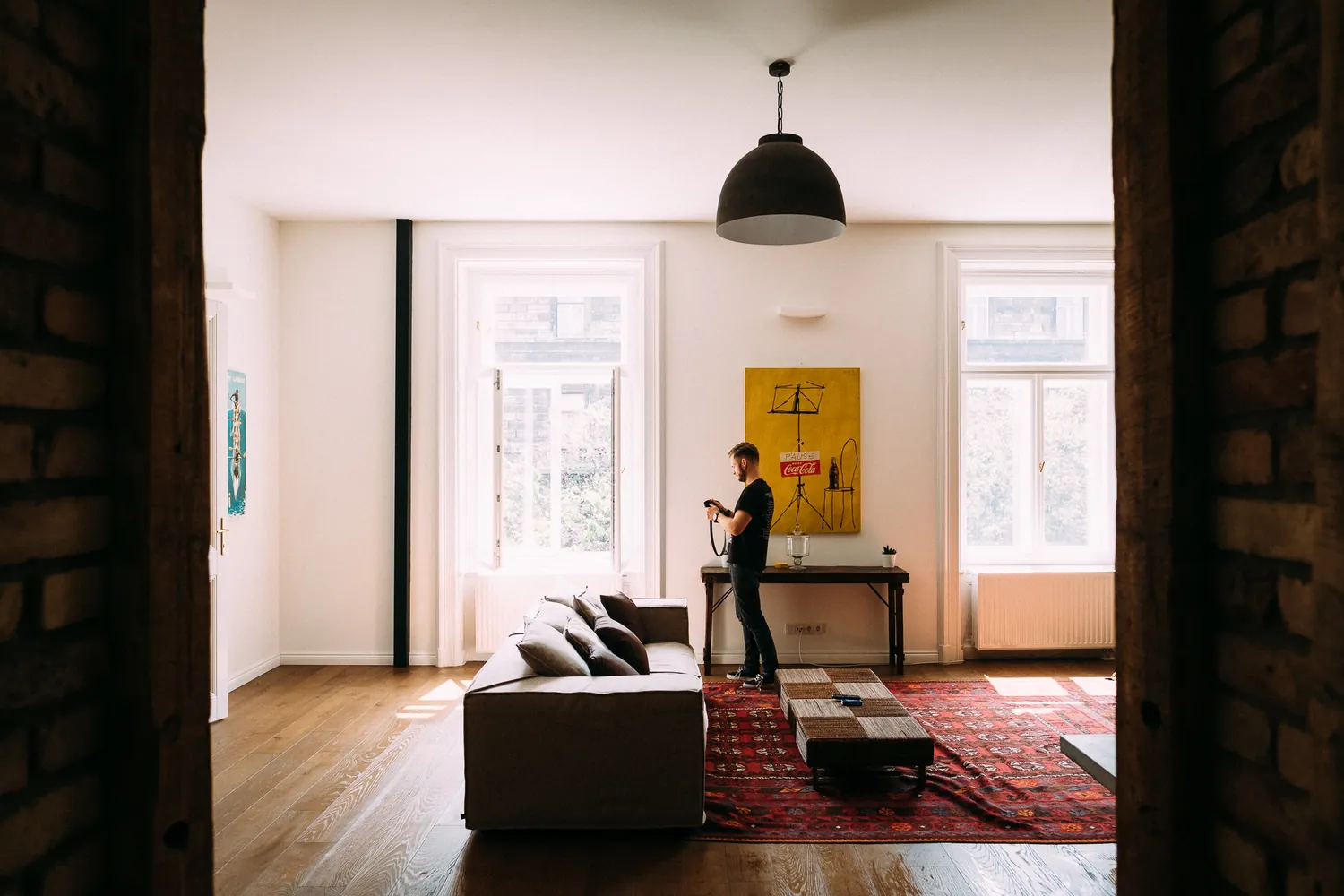 A tall, light apartment room with a dark pendant lamp and a rug