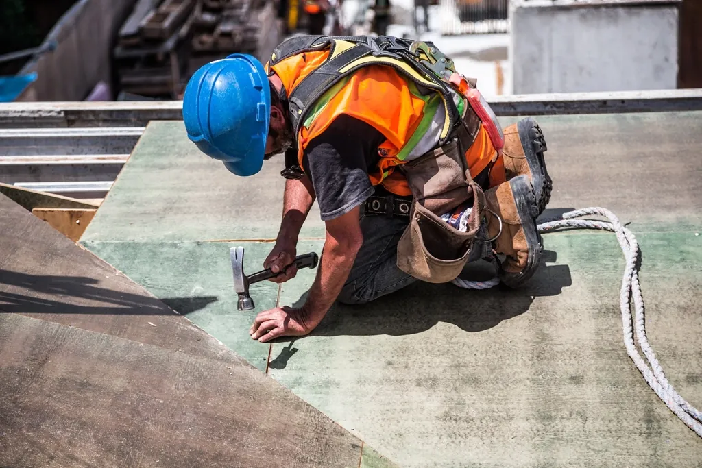 A roofer in a hard hat and hi-vis kneeling on a roof deck, nailing down membrane